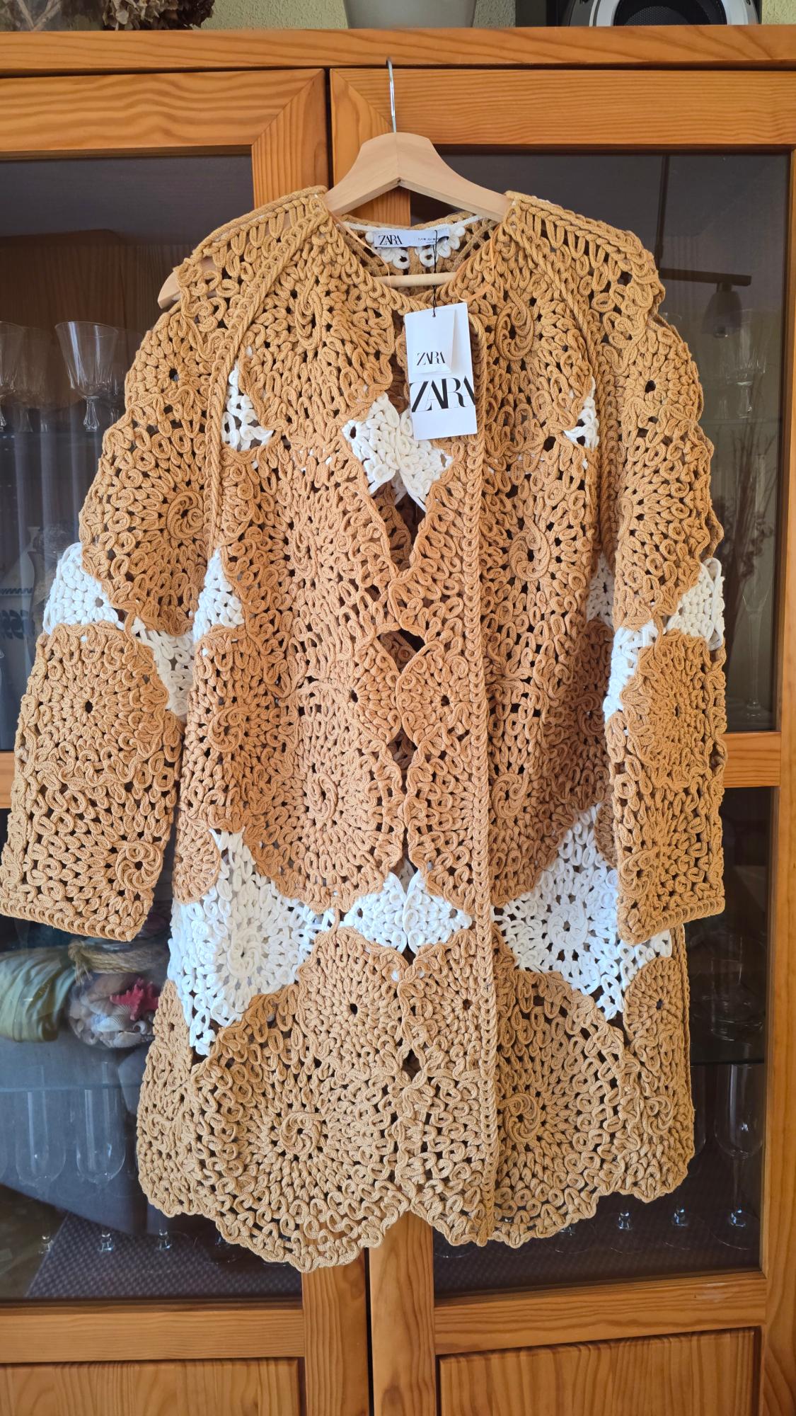 Crochet coat in beige and white tones hanging on a wooden hanger. Visible label with the text "ZARA". Wooden furniture background.