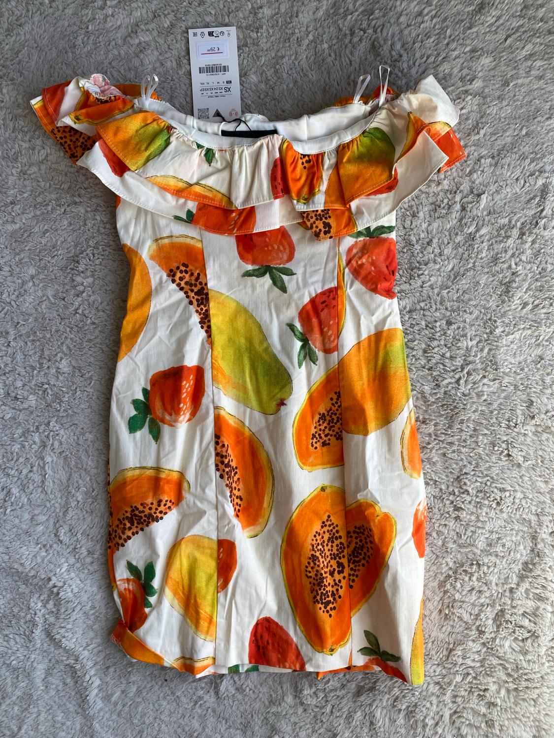 White dress with a papaya and strawberry print, frilled neckline, on a plush rug. Tag with the text €29 and XS.