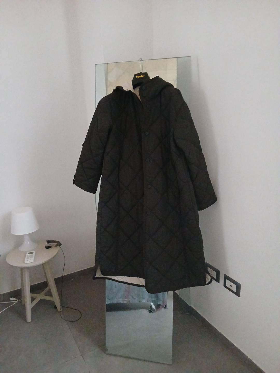 Long dark padded coat hanging in front of a mirror in a room with white walls, next to a small table with a lamp.