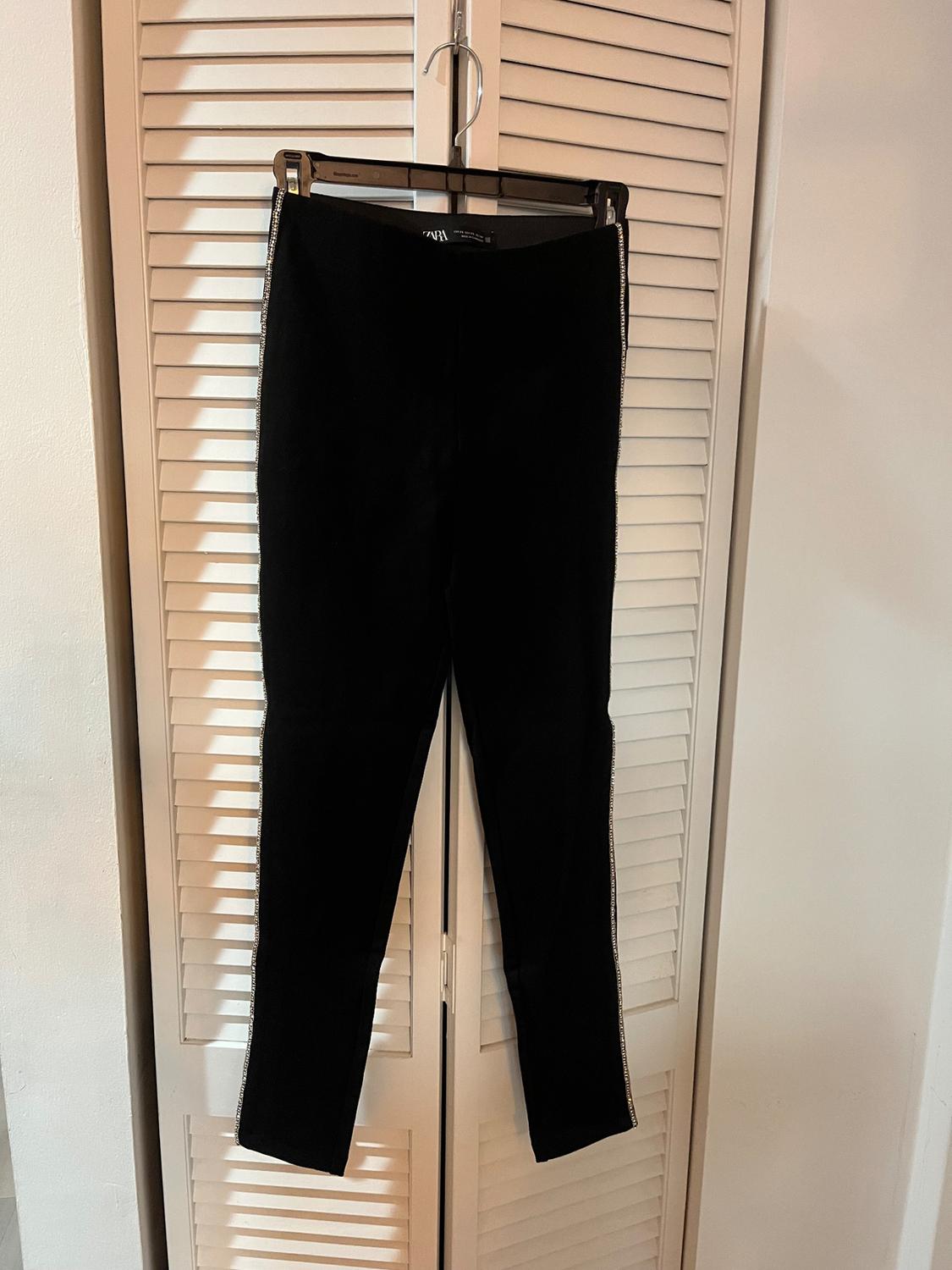 Black trousers hanging on a hanger in front of a white roller shutter door. Shiny ribbon detail on the sides.