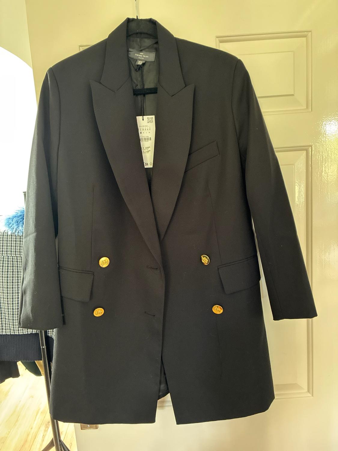 Black double-breasted blazer with gold buttons hanging on a white door. Label with the text ZARA STEFANO PILATI.