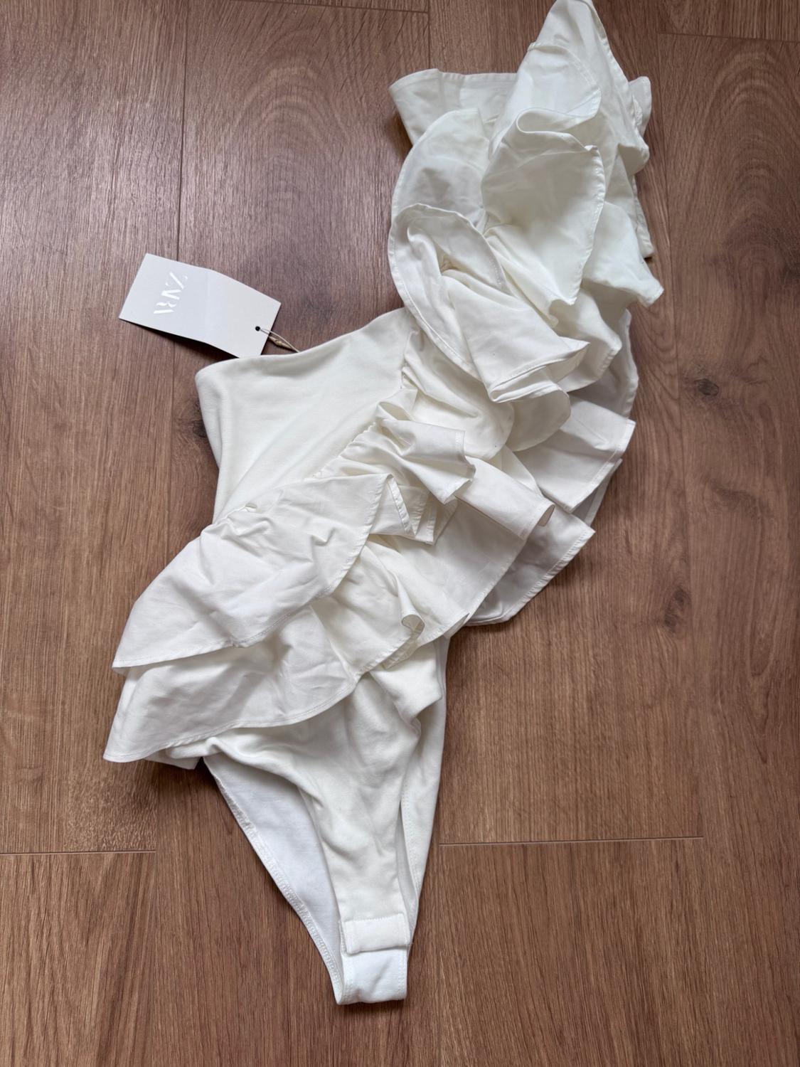 White one-shoulder bodysuit with large ruffles, on a wooden floor. It has a label bearing the text ZARA.