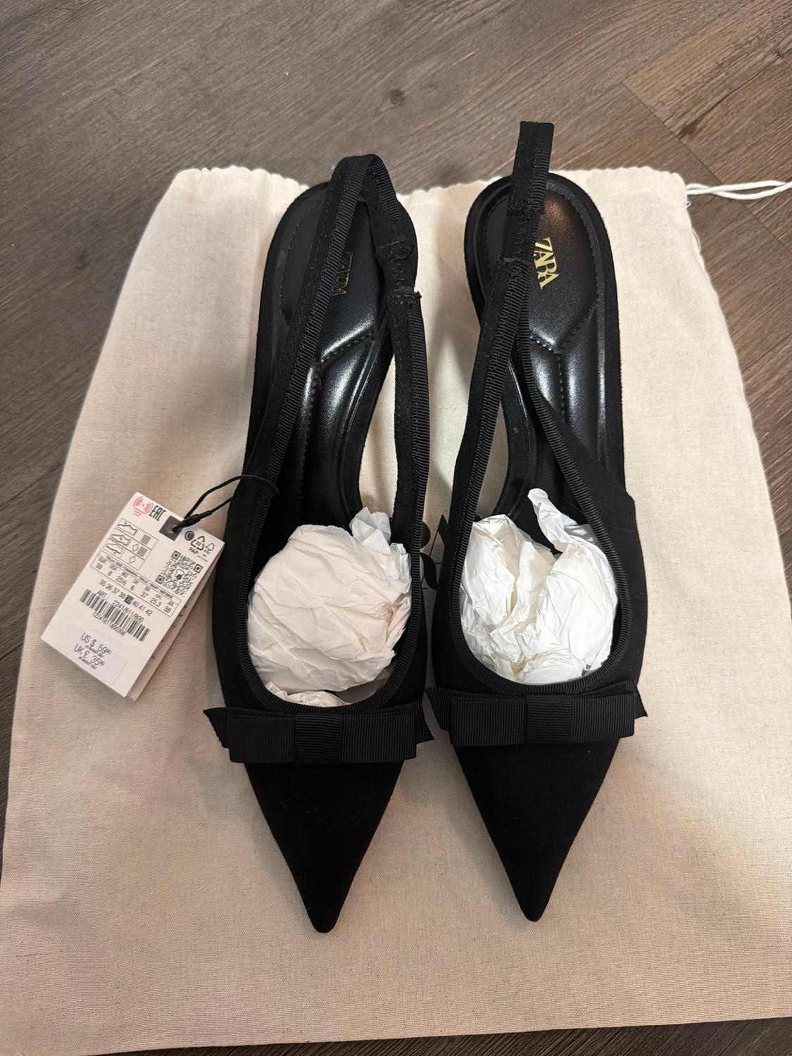 Black high-heeled shoes with a bow, stuffed with paper, on a beige fabric bag. Visible tag with product details.