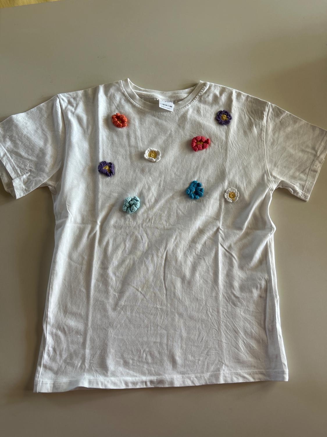 White t-shirt with crochet flowers in various colours: red, blue, purple, white and orange, on a beige surface.