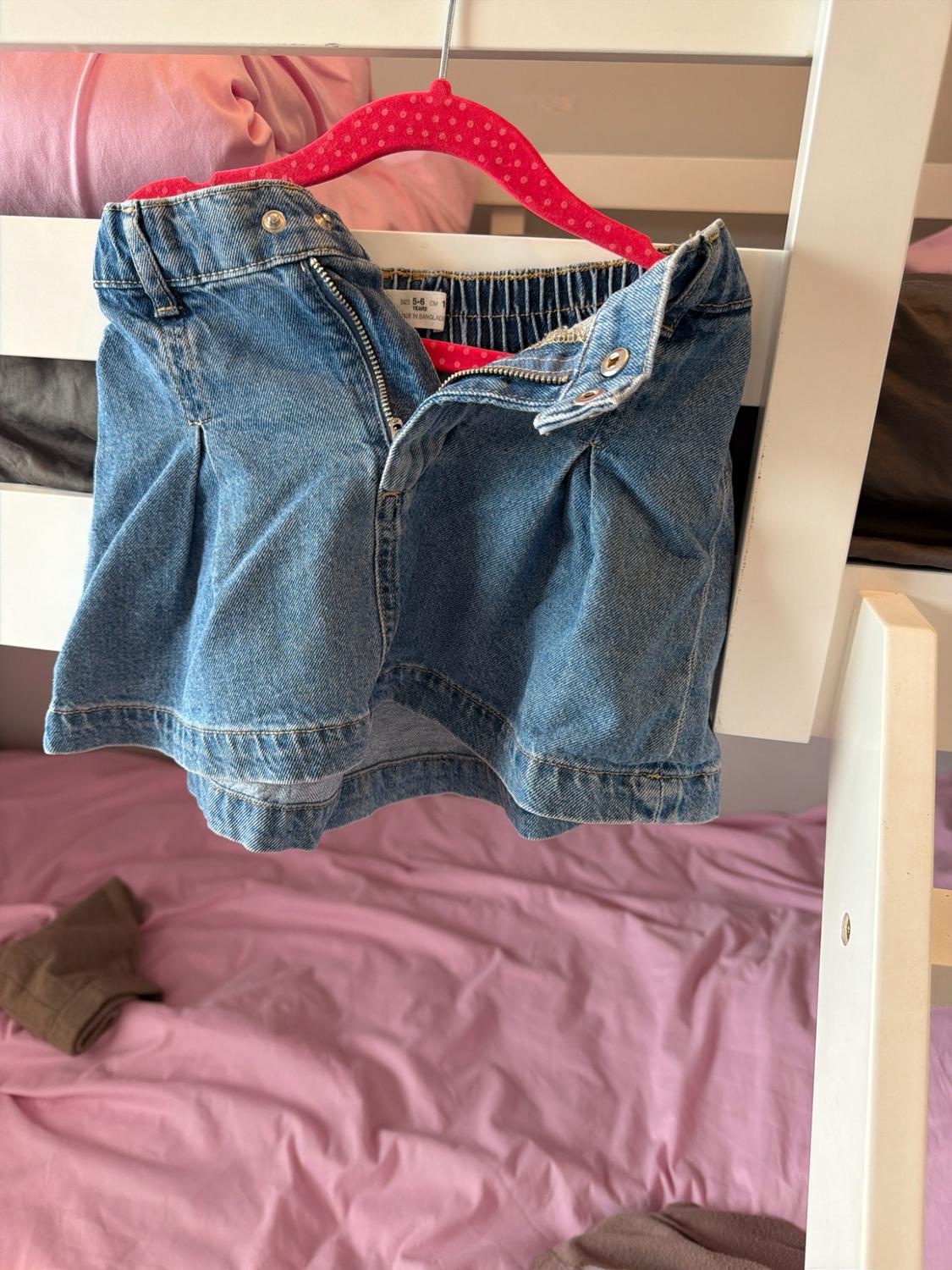 Unbuttoned blue denim shorts on a pink hanger, hanging from a white bed frame. Against a background of pink bed sheets.