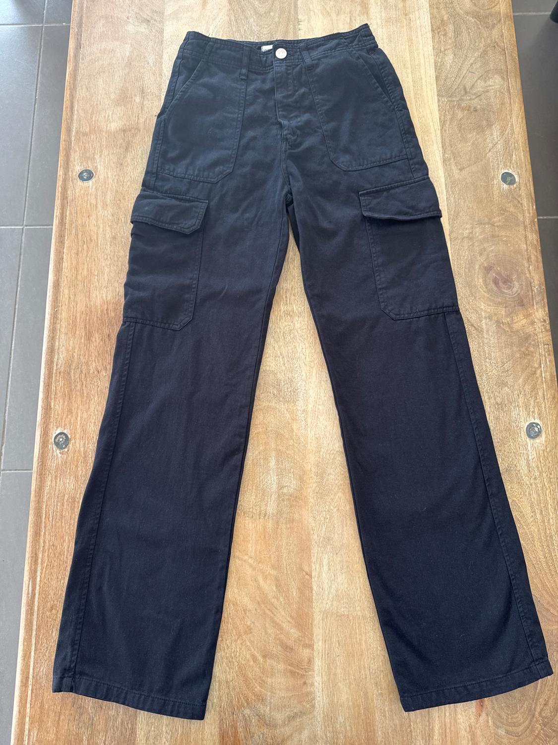 Black cargo trousers with side pockets, placed on a light wooden table.