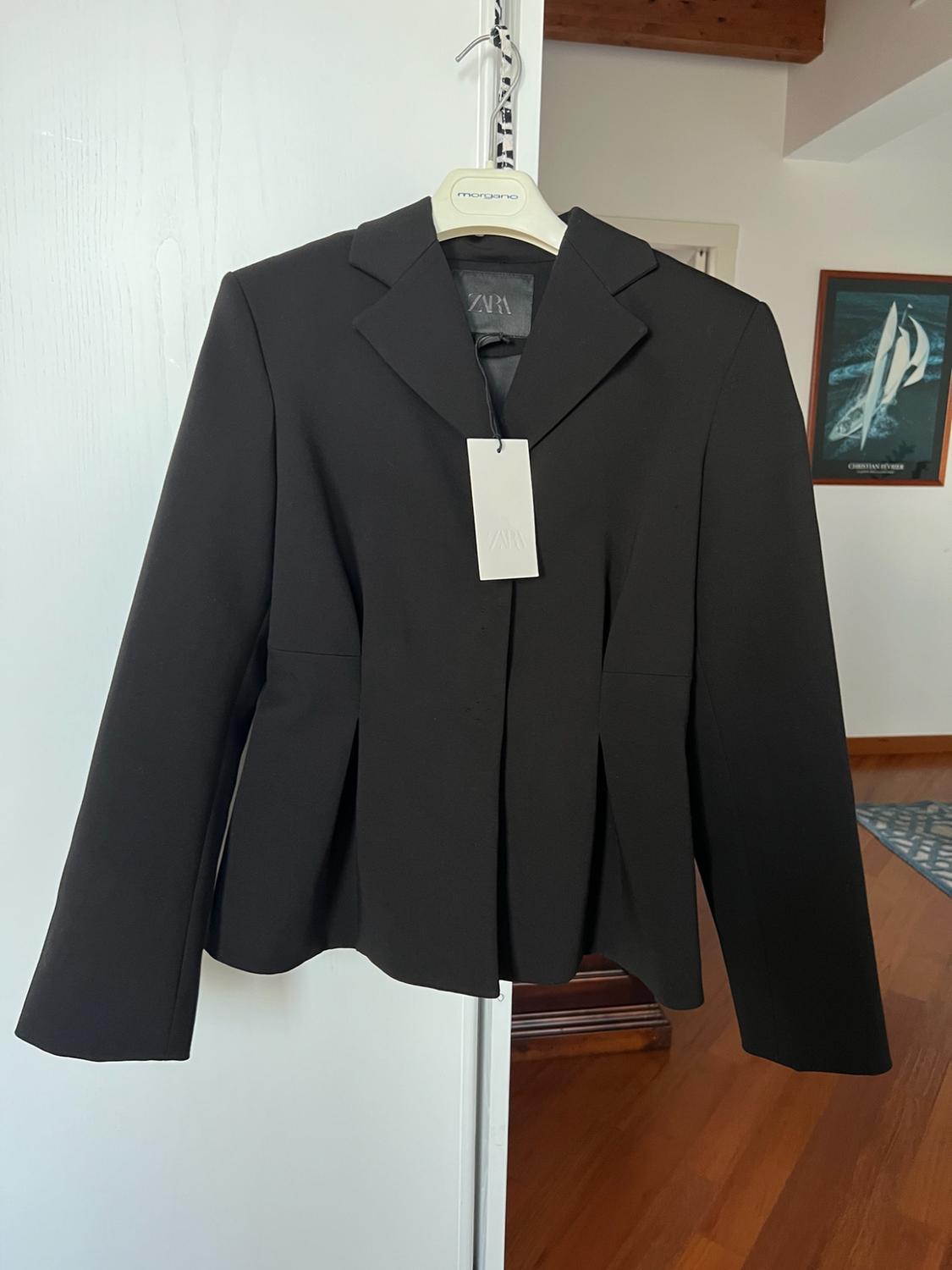 Black pleated jacket hanging on a door. Text: ZARA, morgano, CHRISTIAN FEVRIER. Background with wooden flooring and a sailing boat poster.