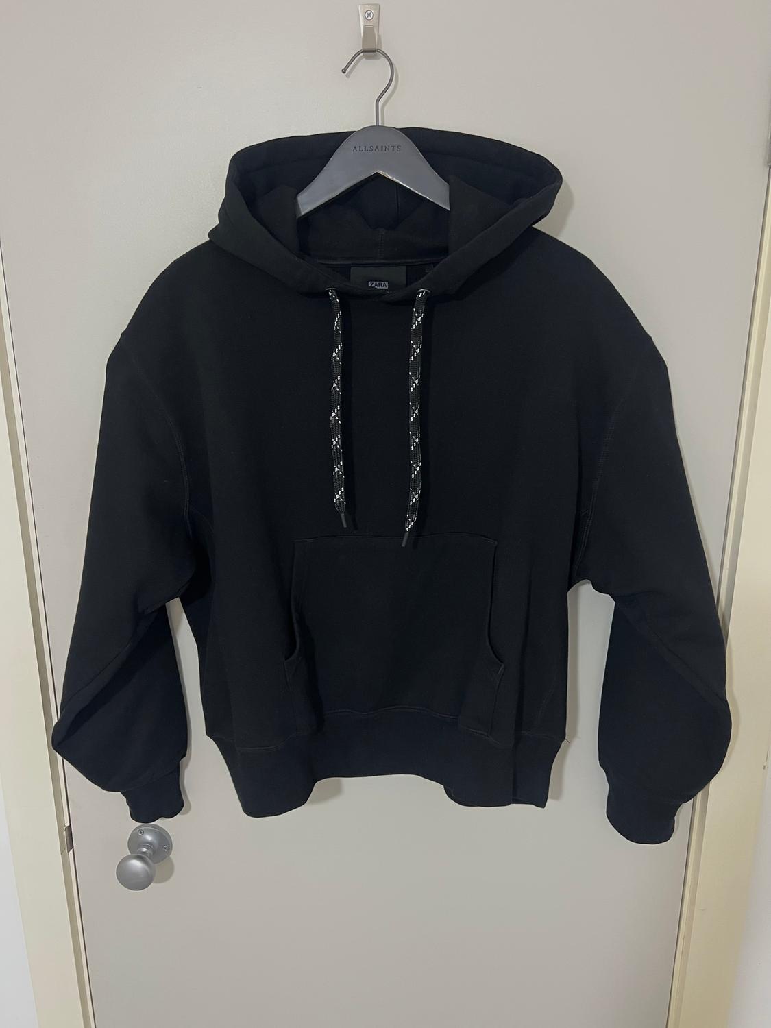 Black hoodie with drawstrings hanging on an AllSaints hanger over a white door. Front pocket visible.
