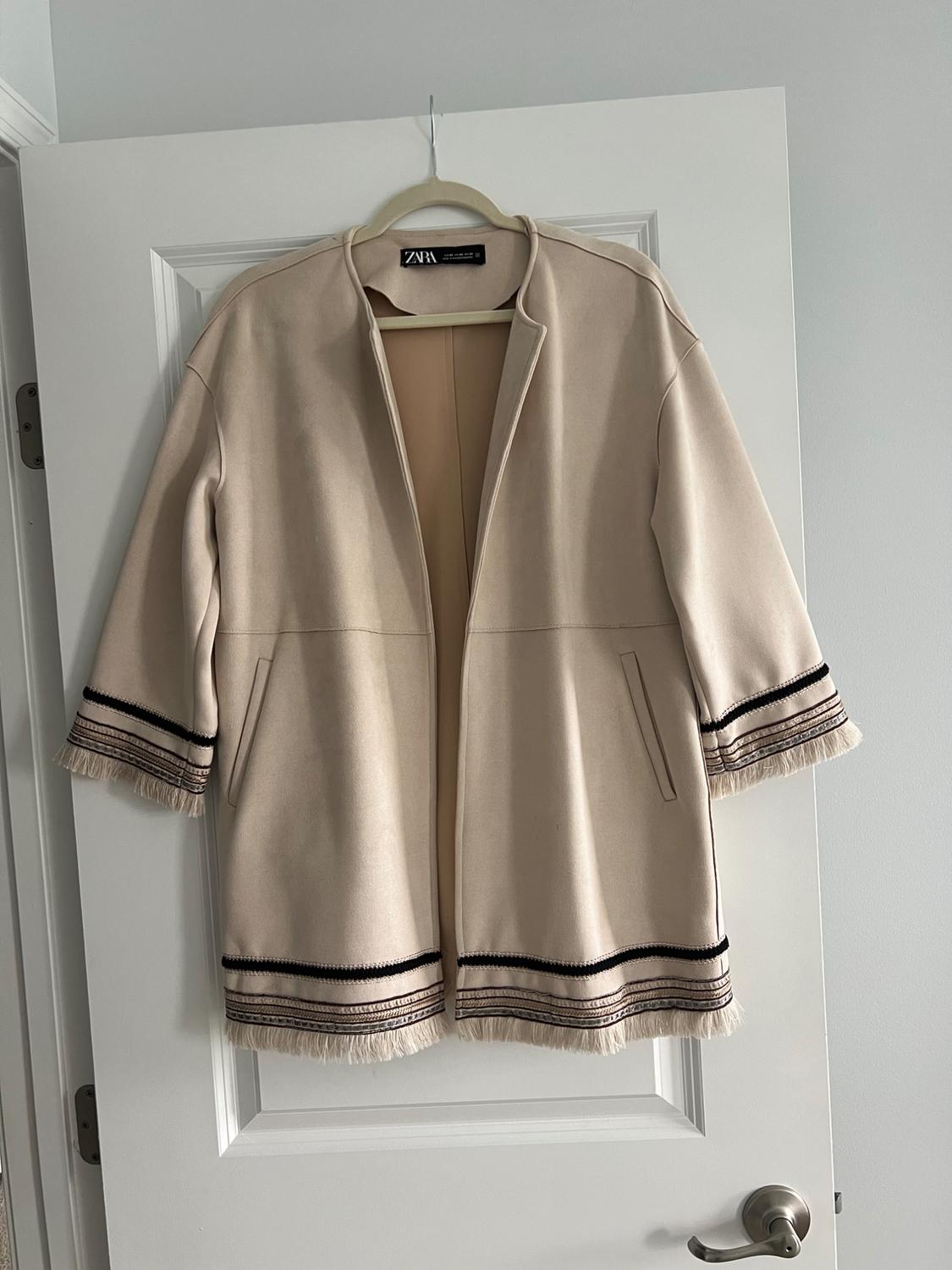 Beige jacket with fringe and black details hanging on a white door. Visible label: "ZARA". Minimalist and modern setting.