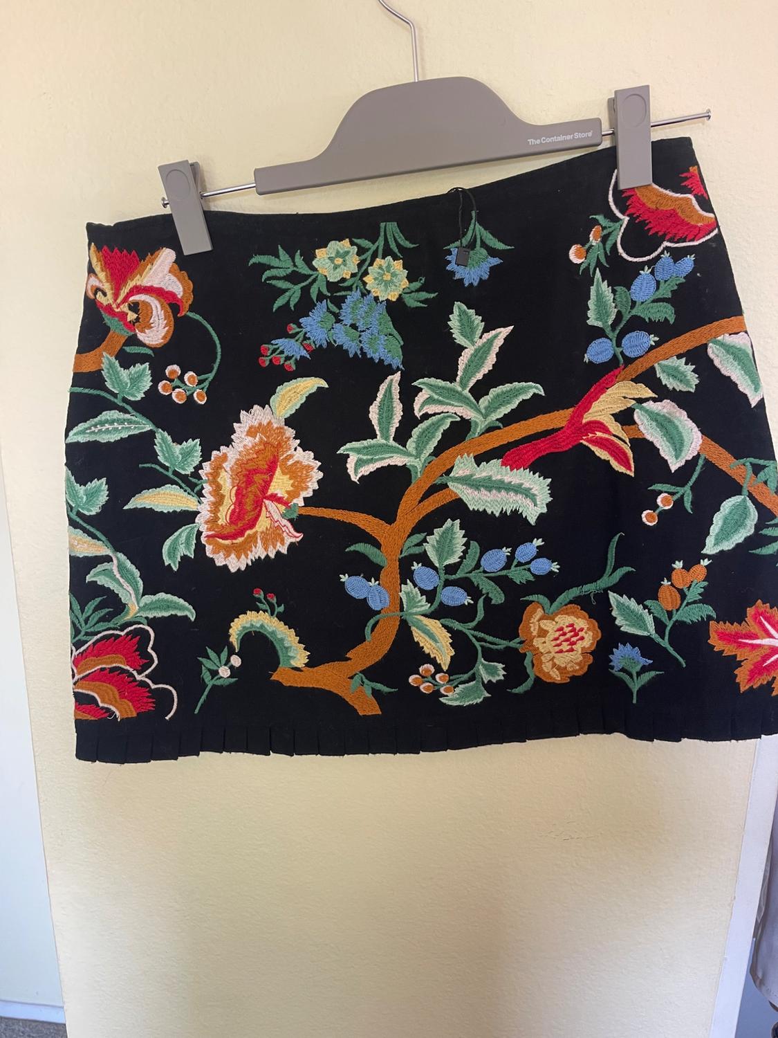 Black skirt with colourful floral embroidery, hanging on a grey hanger. Beige wall background.