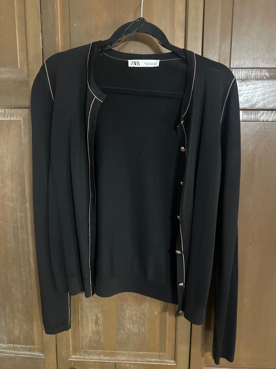 Black knitted jacket with light trim and buttons, hanging on a hanger in front of a wooden door. Visible label: ZARA.