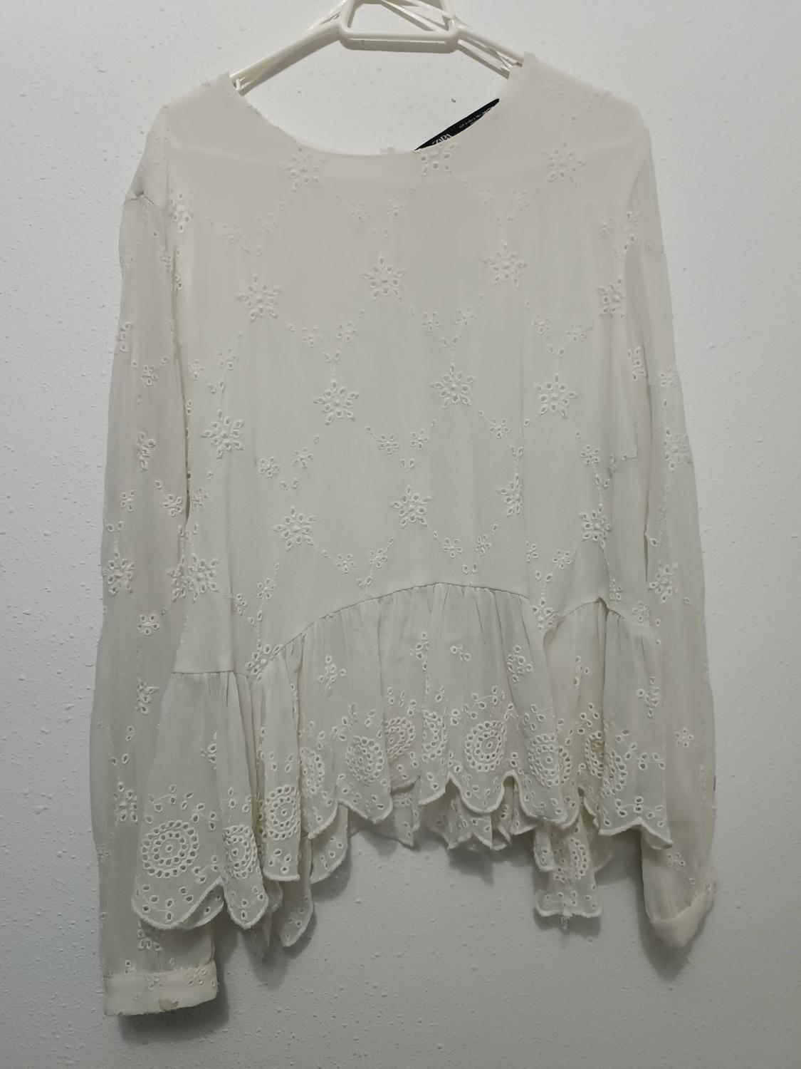 White lace blouse hanging on a hanger, with embroidered floral details and ruffles on the bottom edge, on a white background.