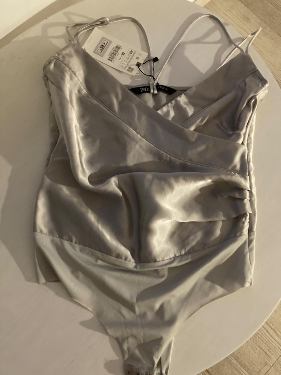 Grey satin bodysuit with thin straps on a white table. Visible labels showing price and size. Warm light illuminates the scene.