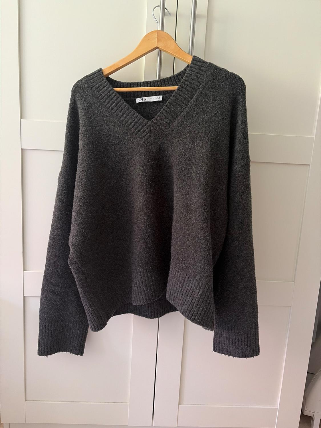 Dark grey chunky knit V-neck jumper, hanging on a wooden hanger in front of a white wardrobe.
