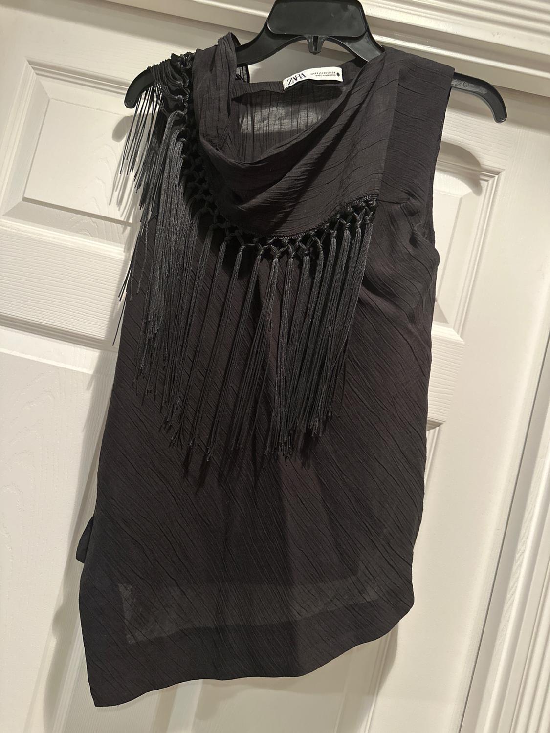 Sleeveless black blouse with fringe hanging from the neck, hung on a black hanger in front of a white door. Visible label: ZNX.