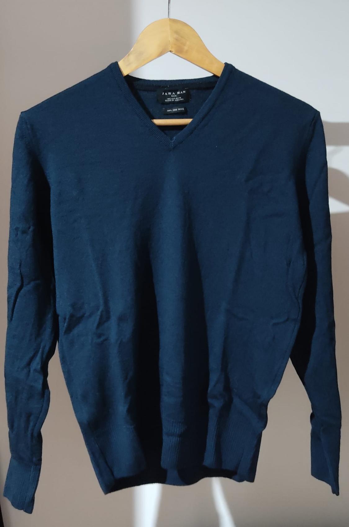Μπλε σκούρο πουλόβερ με V-λαιμό, κρεμασμένο σε ξύλινη κρεμάστρα, μπροστά από ανοιχτόχρωμο φόντο. Ορατό κείμενο: ZARA MAN.