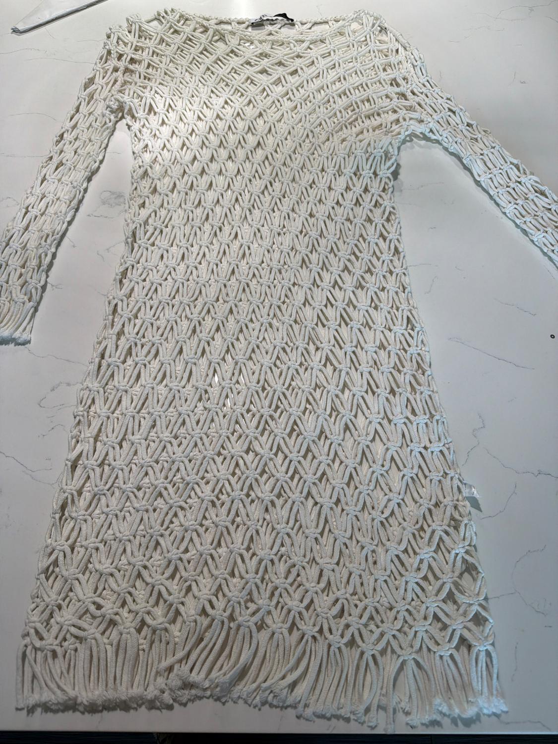White long-sleeved dress with macramé-style knot detailing and fringe at the hem, laid out on a light marble surface.