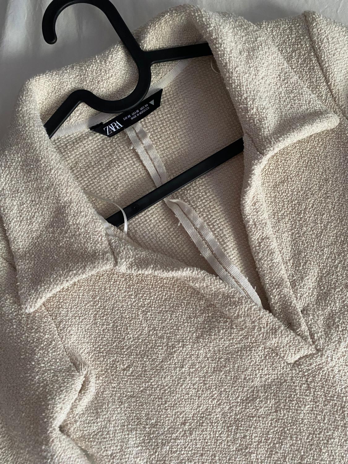 Close-up of a cream garment with a collar and textured fabric on a black hanger. Text: ZARA EUR XS USA XS MEX 24 MADE IN MOROCCO.