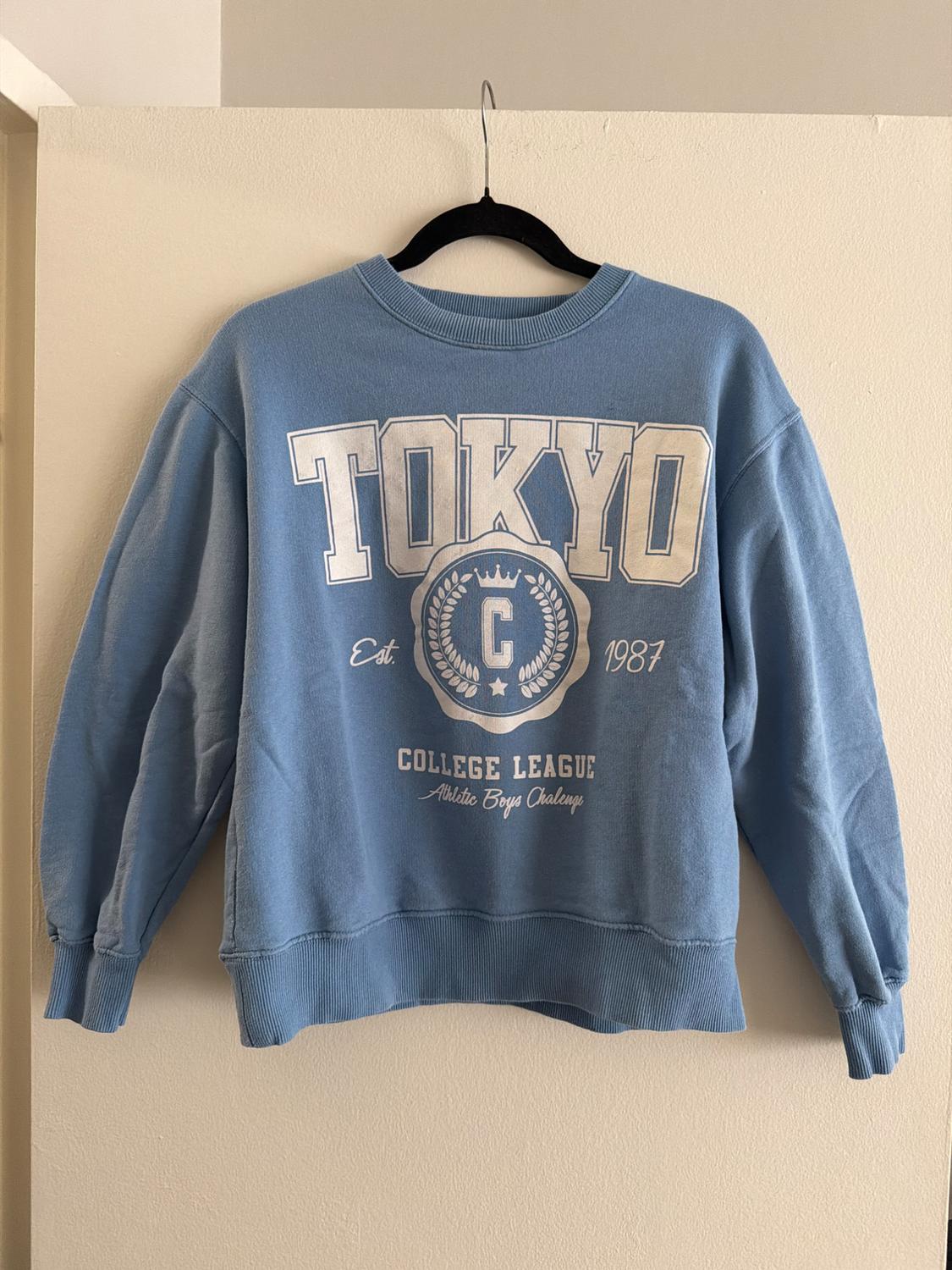 Blue sweatshirt hanging on a black hanger, with "TOKYO", "Est. 1987", "COLLEGE LEAGUE" and "Athletic Boys Club" text in white.