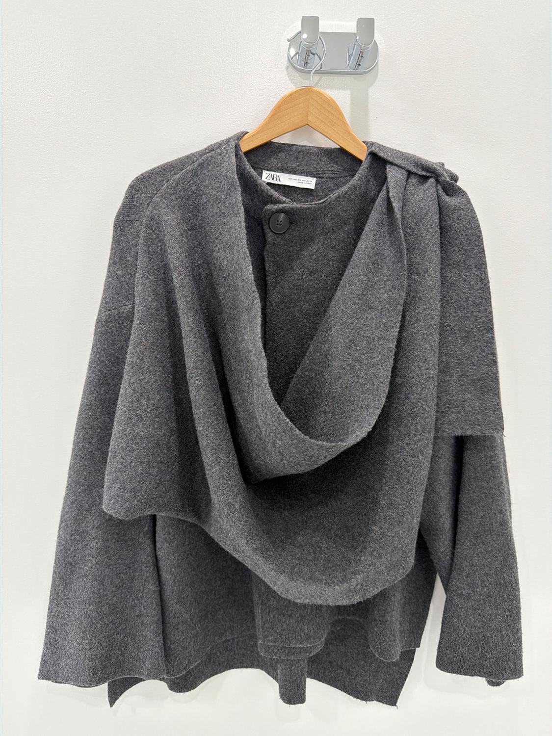 Gray knit sweater with a draped neckline and one button, hanging on a wooden hanger against a white background. ZARA is visible on the label.