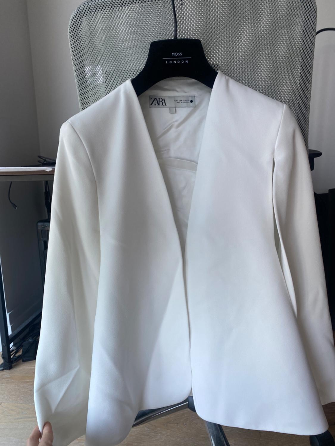 White jacket with a ZARA label on a black hanger with MOSS LONDON text, hung on an office chair.