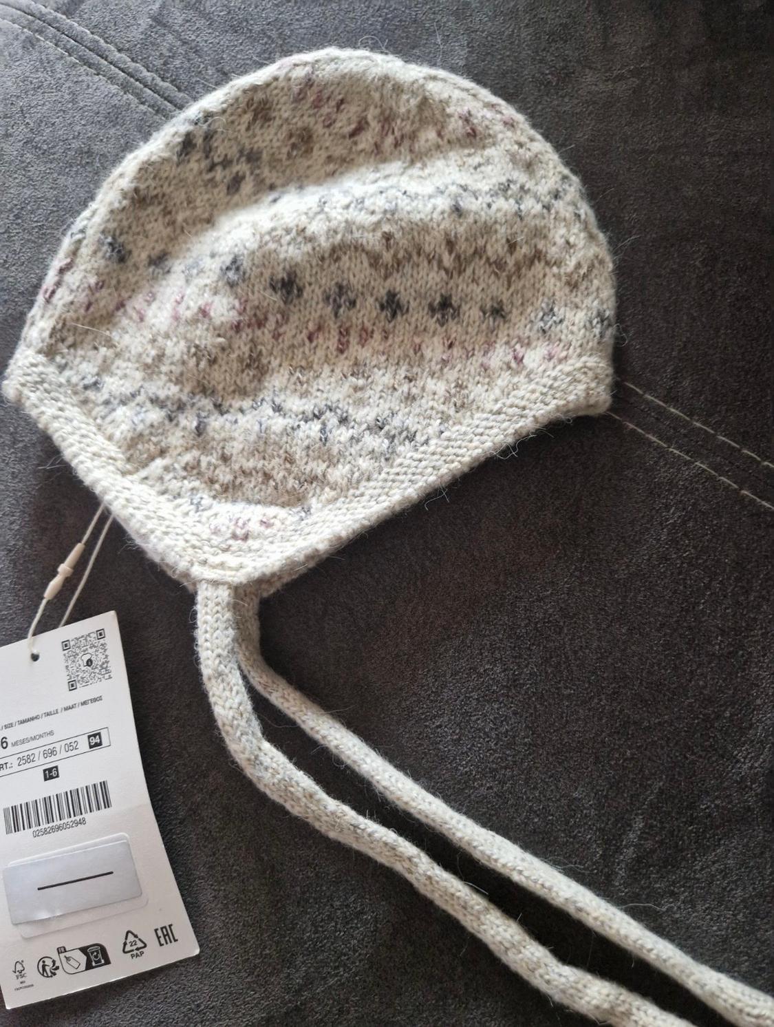 Woolly knitted hat in beige tones with a dark dot pattern and product label on a grey background.