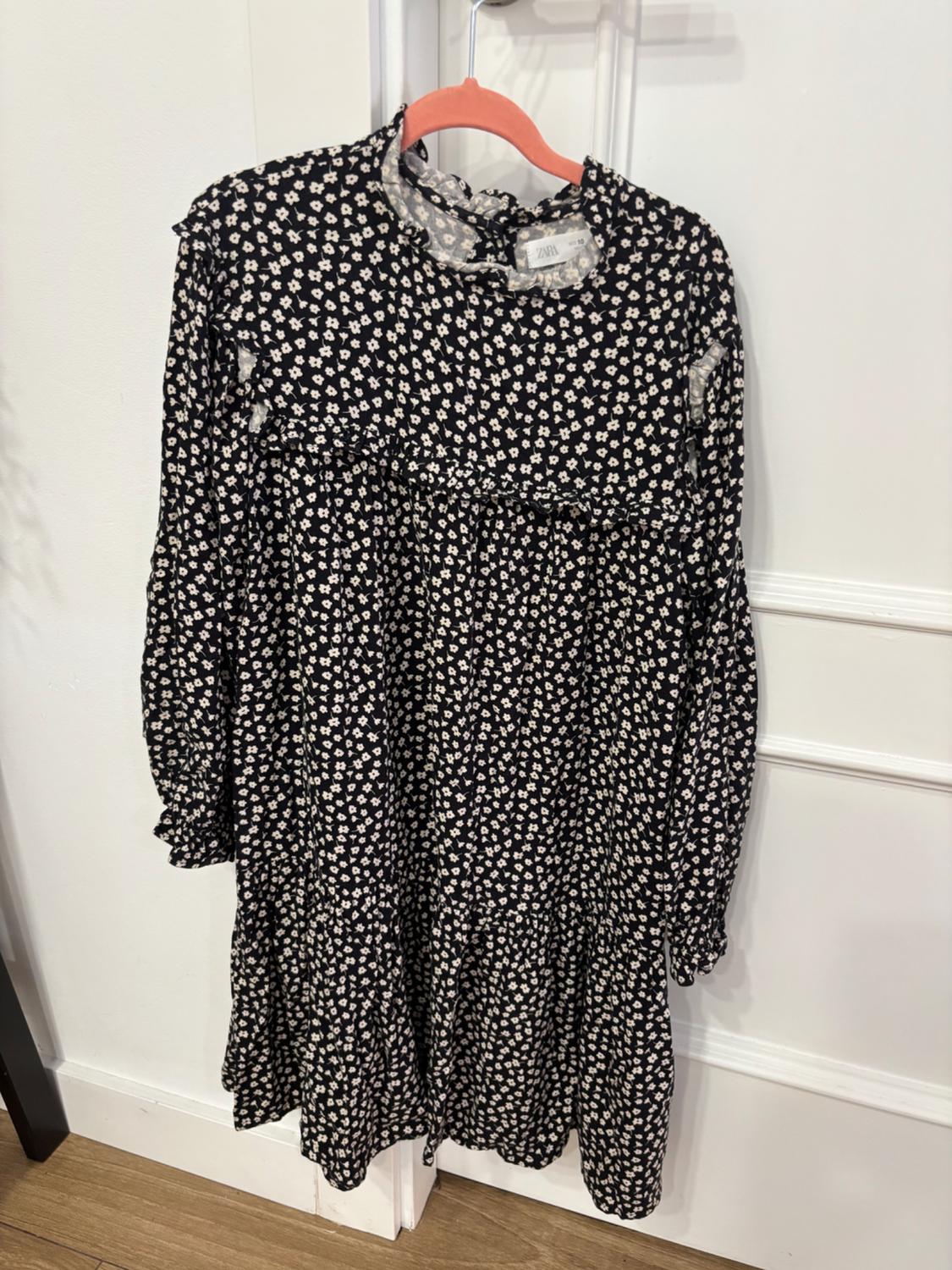 Black dress with a small white floral print, hanging on a red hanger over a white door. Label visible.