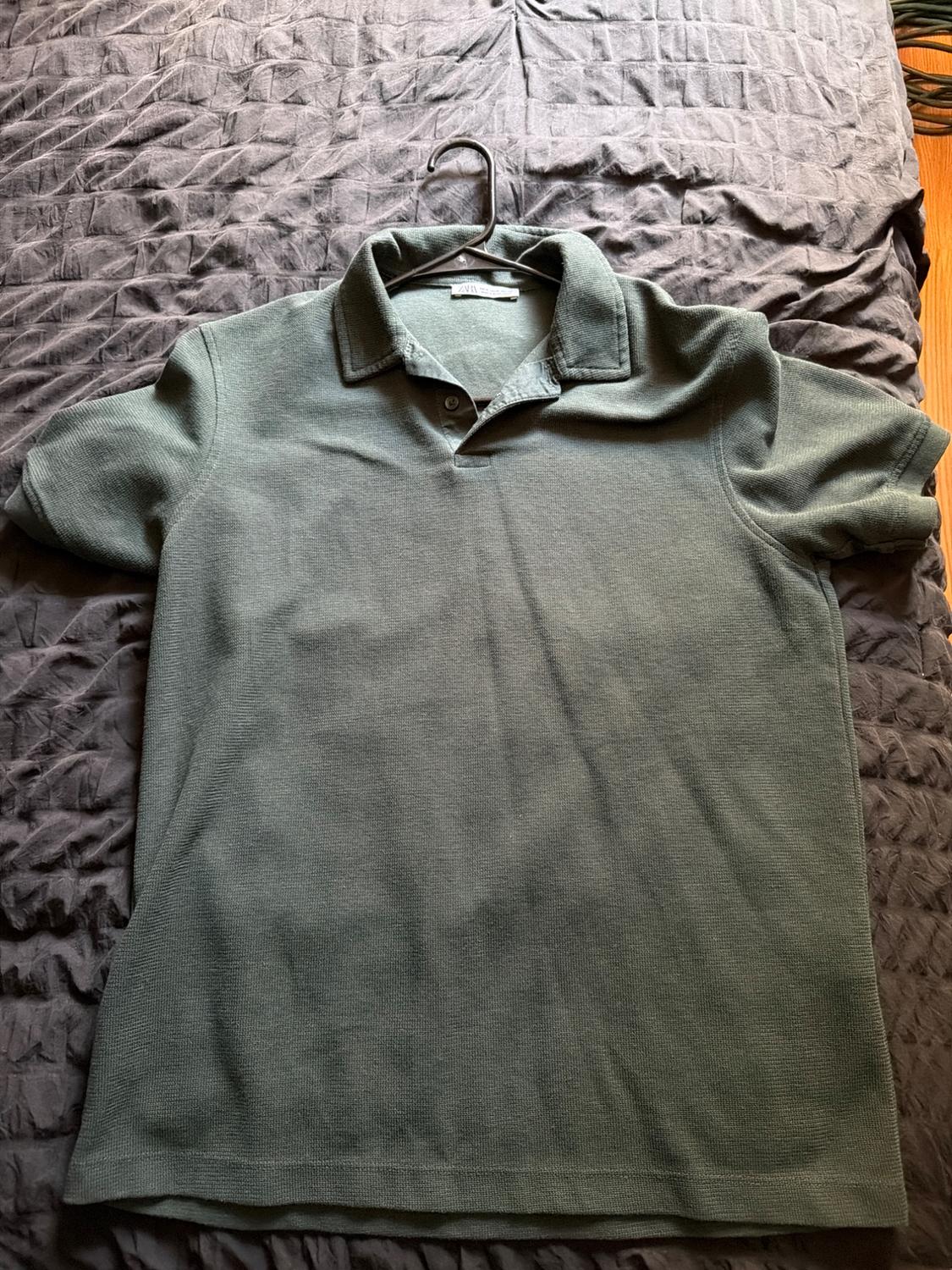 Short-sleeved green polo shirt with collar and buttons, placed on a hanger over a dark wrinkled fabric surface.