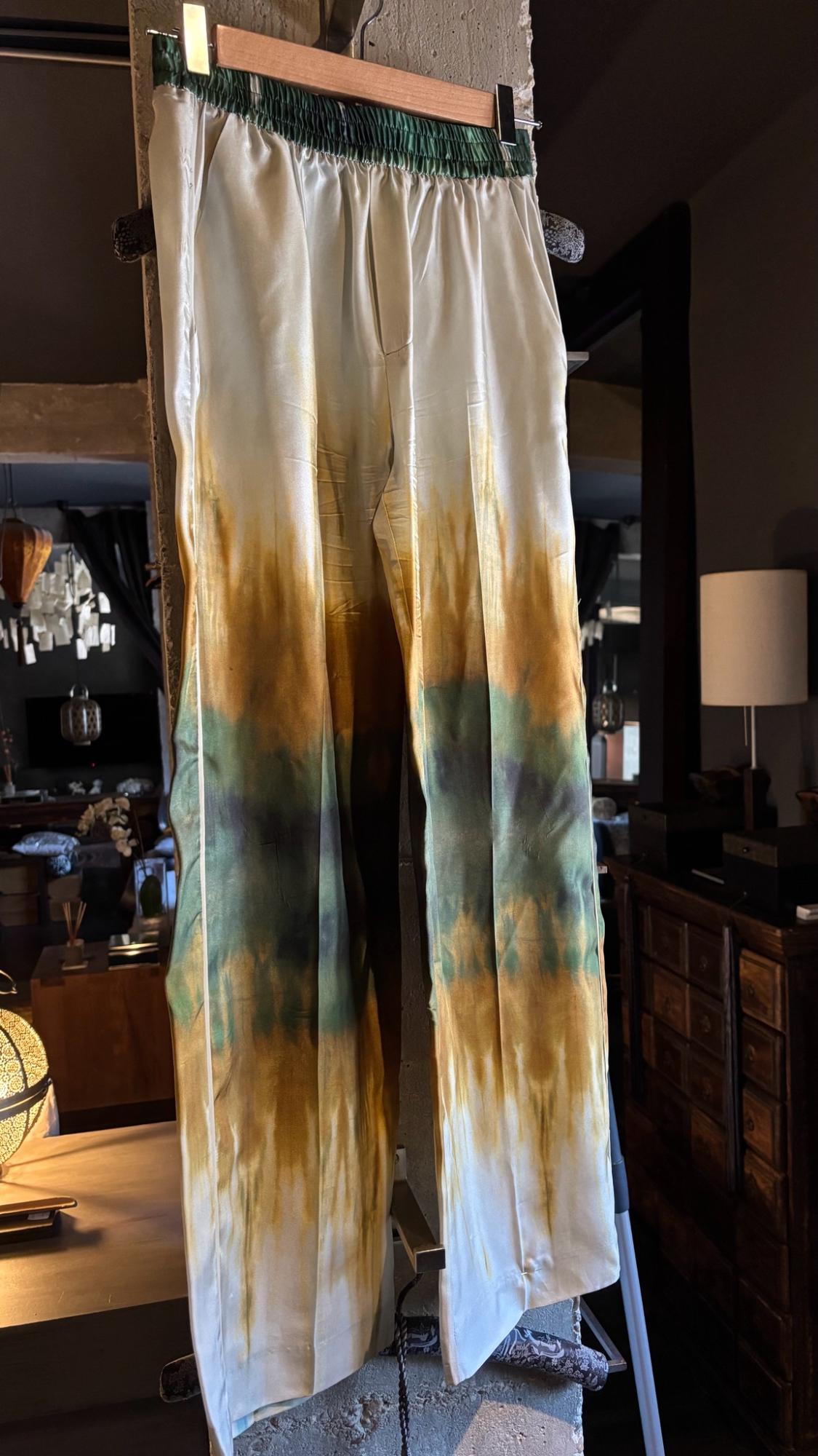Satin trousers with an elasticated waist and an ombré print in shades of white, ochre and green, hanging on a hanger inside an elegant living room.