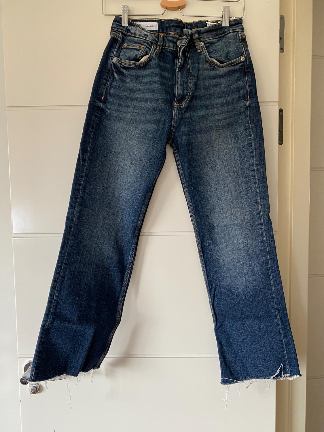 Worn blue jeans hanging on a white door, with frayed hem and five visible pockets.