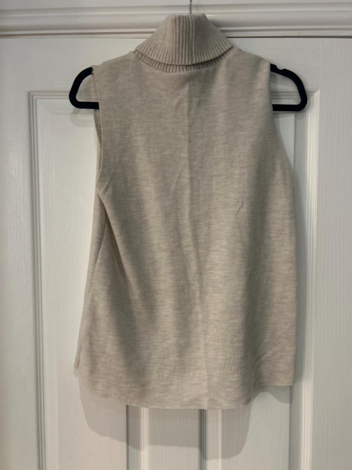 Beige sleeveless jumper with a ribbed roll neck, hanging on a black hanger over a white door.