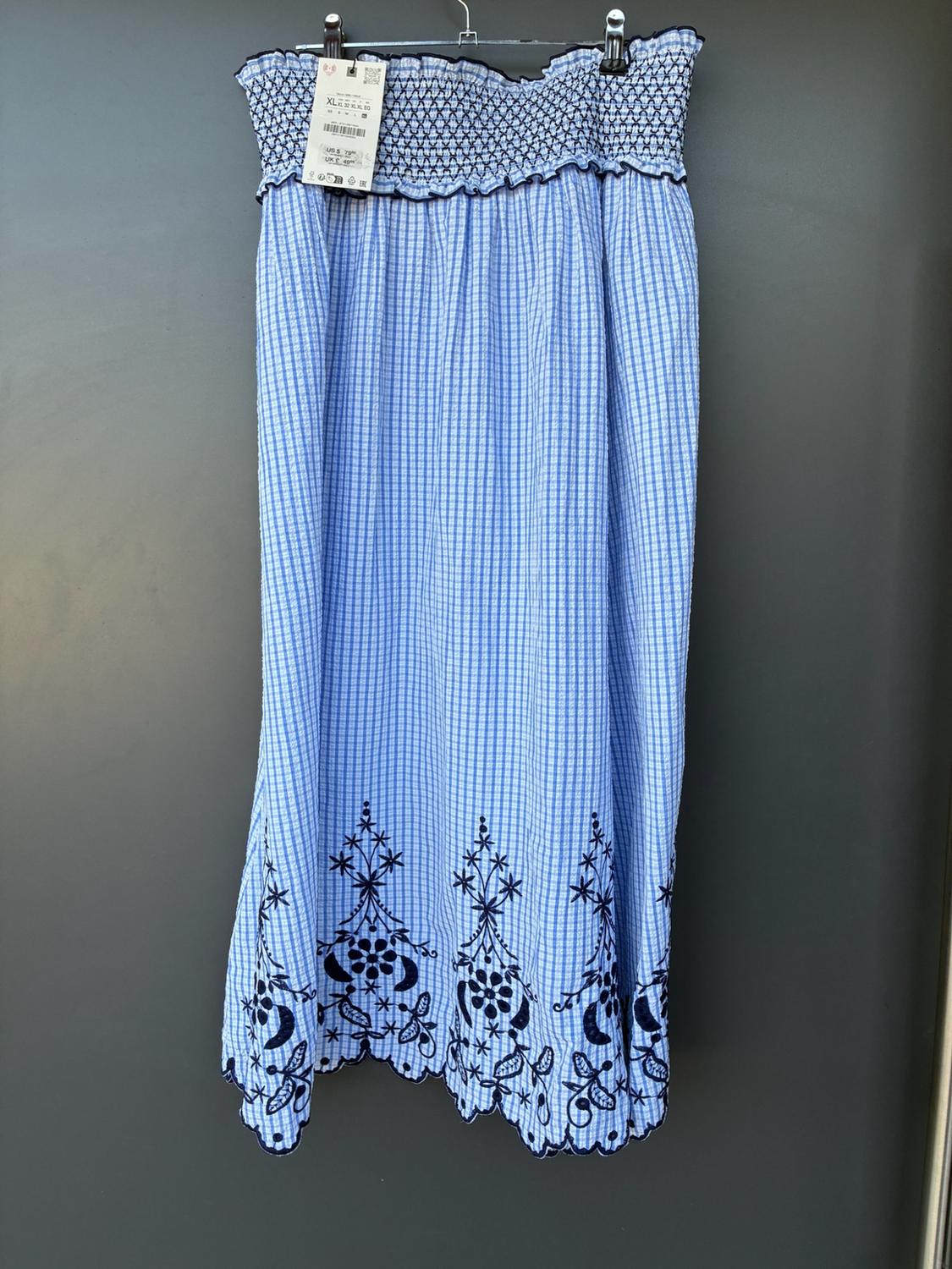 Blue and white checked dress on a hanger. Its gathered neckline and scalloped hem with dark blue floral embroidery stand out.