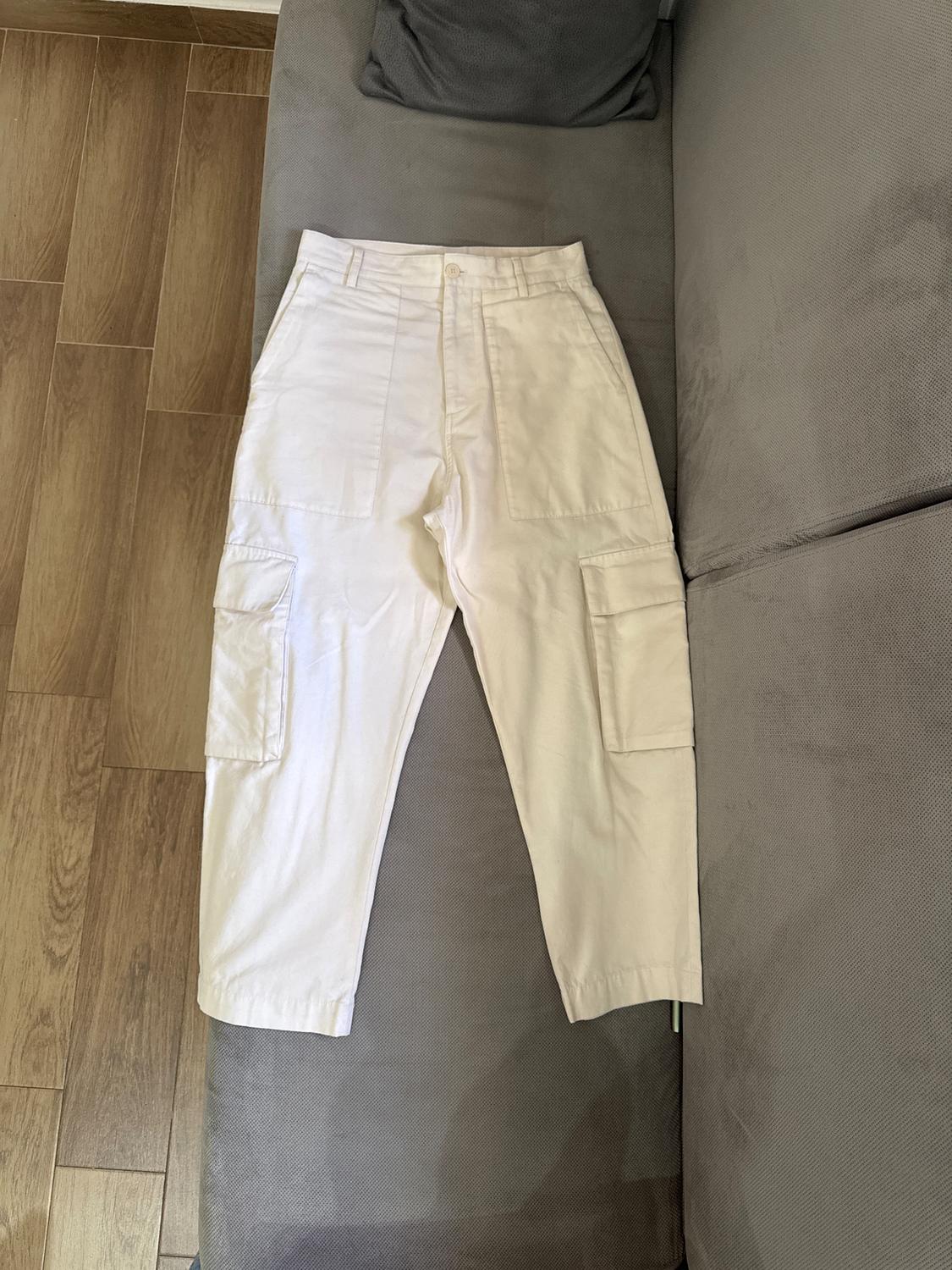 White cargo trousers on a grey sofa, with side pockets and a button at the waist. Wooden floor visible next to it.