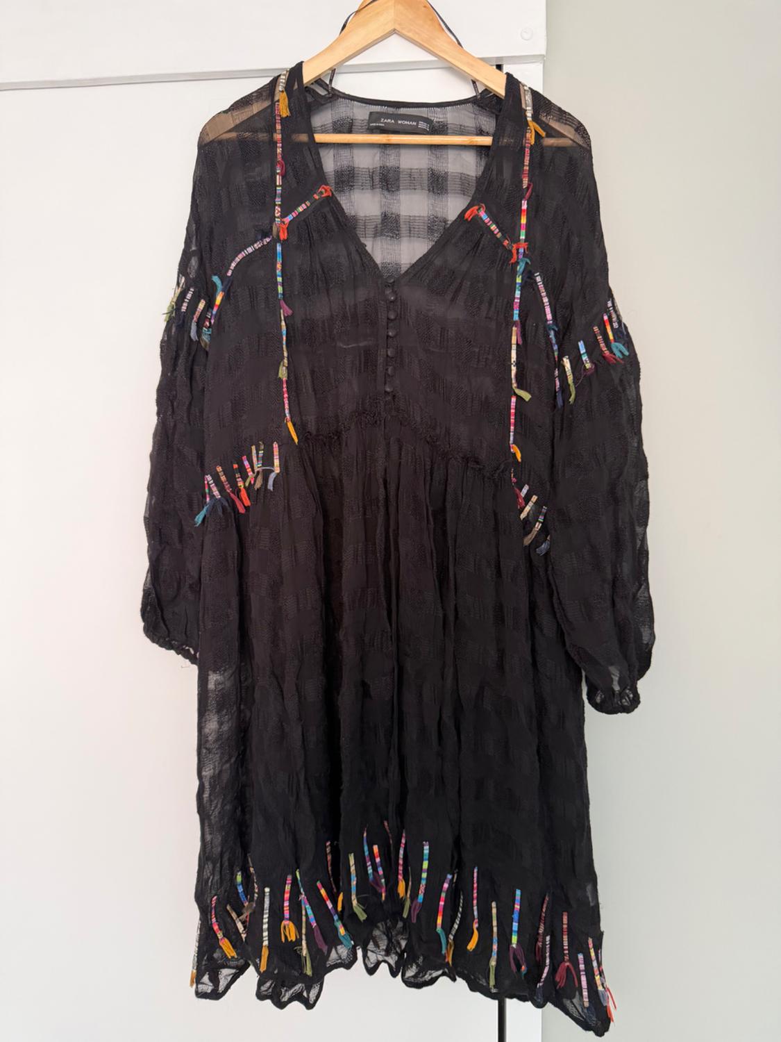 Semi-sheer black dress with colourful ribbon detailing and front buttons, hanging on a hanger. Text: ZARA WOMAN.