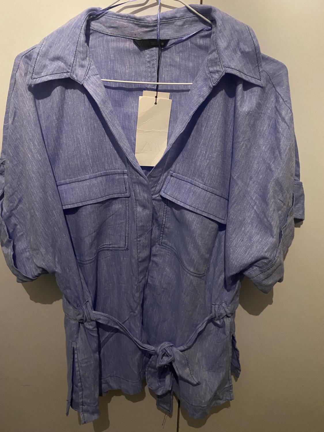 Light blue shirt with front pockets and a knotted belt, hanging on a hanger. Visible label with text "ZARA".