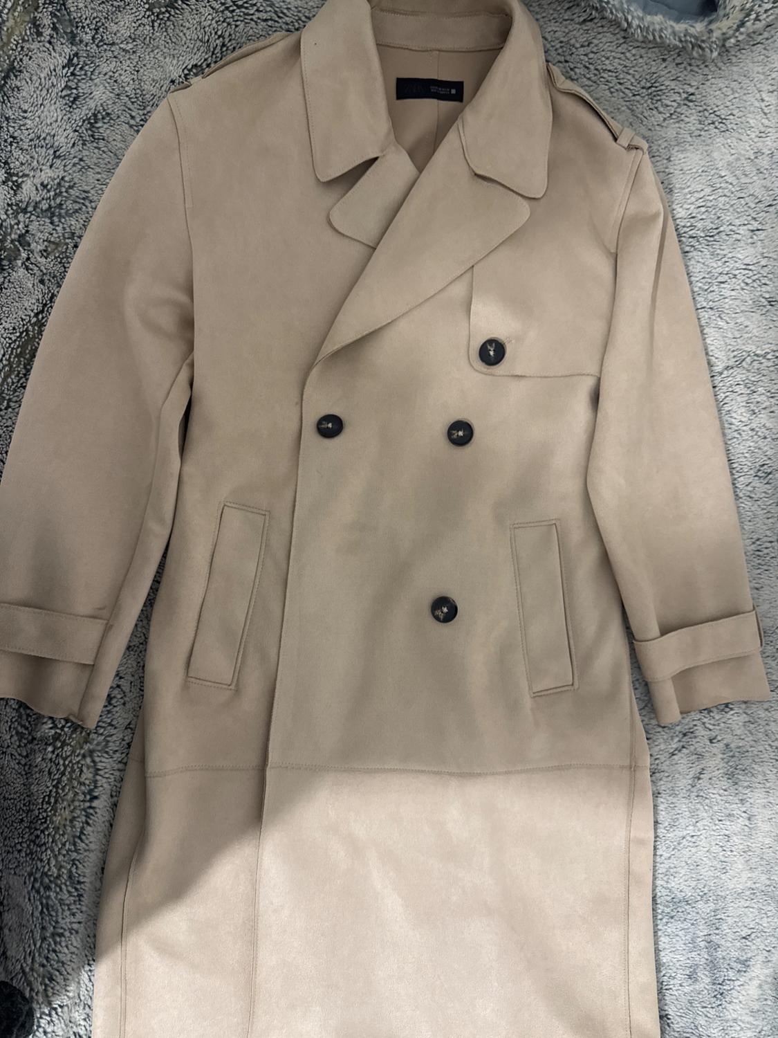Beige double-breasted coat on a grey textured surface. Visible label on the inside of the collar.