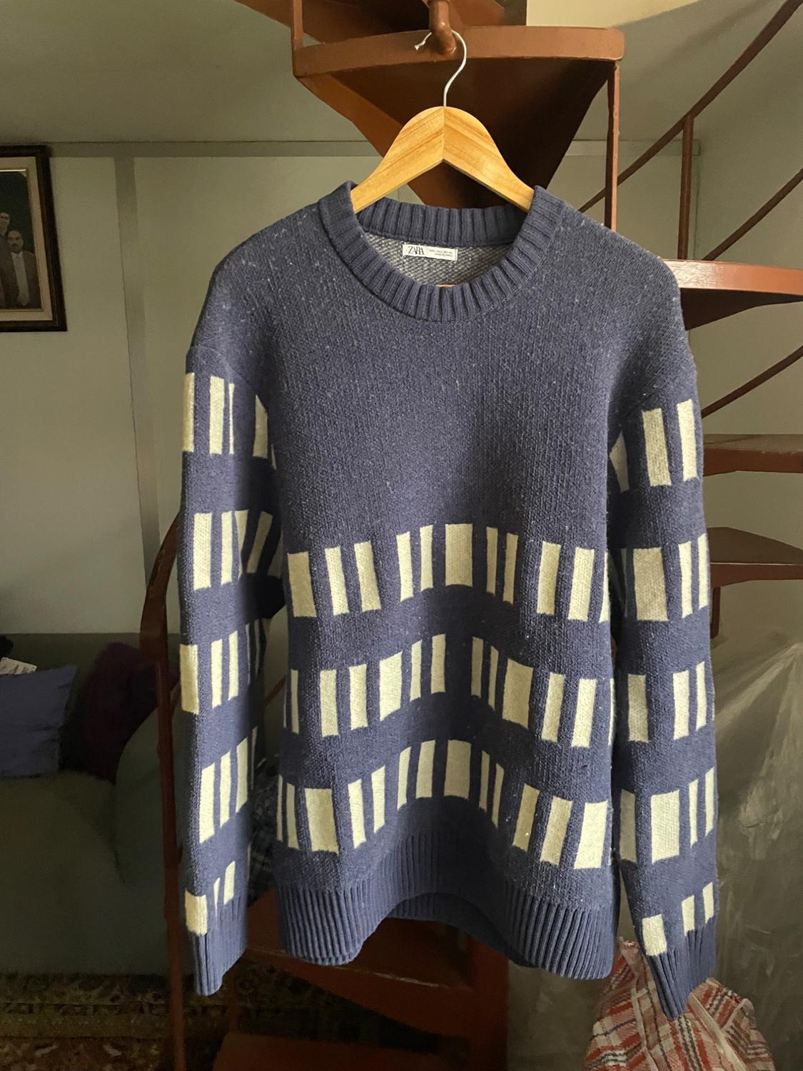 Blue jumper with a white geometric pattern, hanging on a wooden hanger in front of a spiral staircase in an indoor setting.