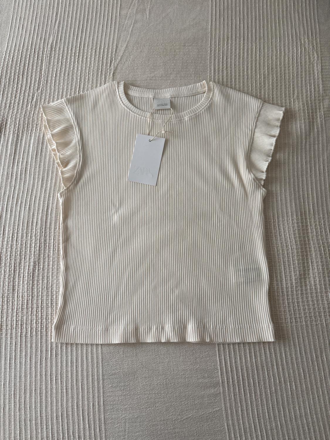 White ribbed T-shirt with short, ruffled sleeves, on a textured surface. Visible hang tag.
