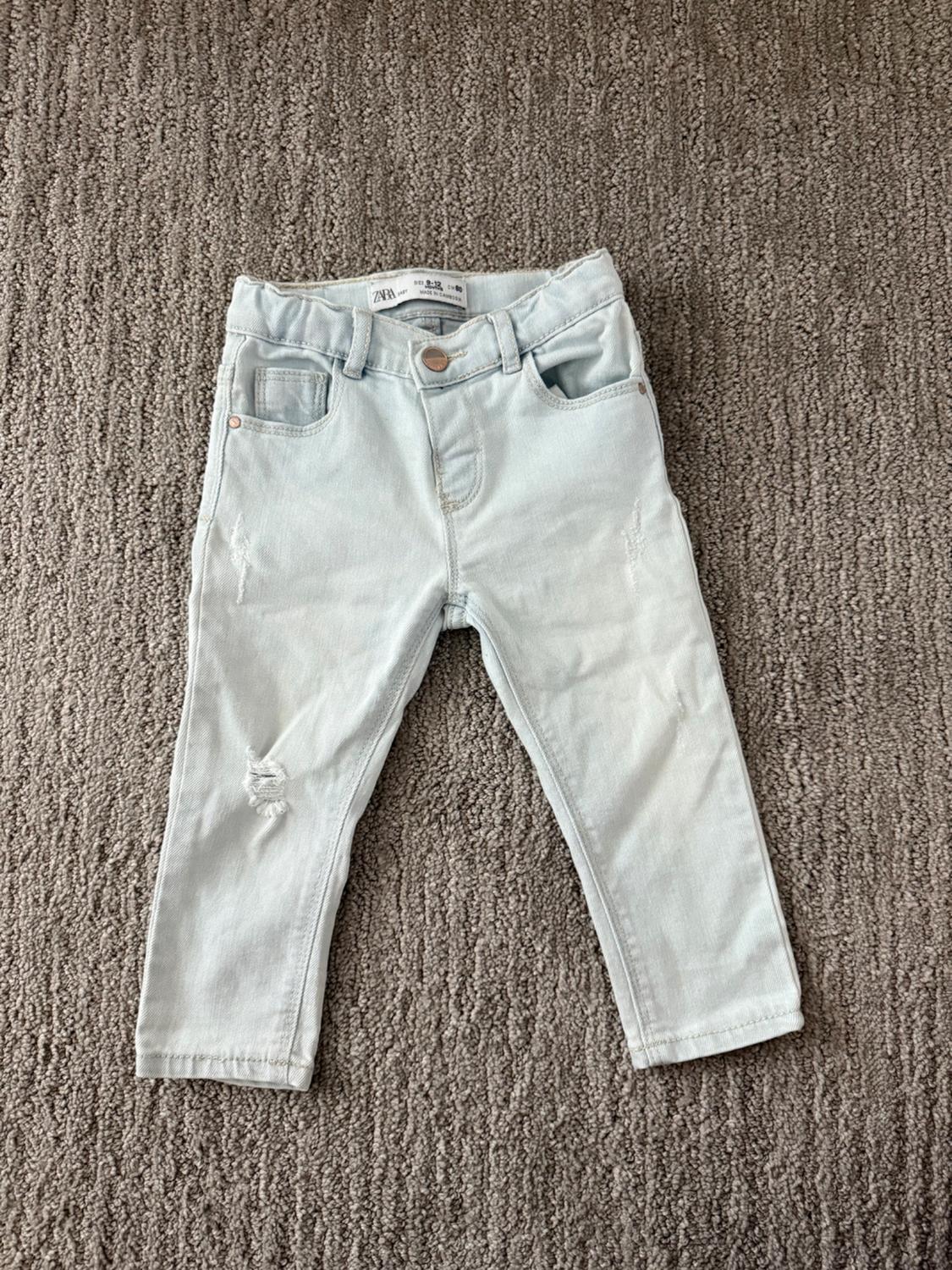 Light-wash denim jeans for kids on a grey rug, featuring distressed detailing and a visible label on the waistband.