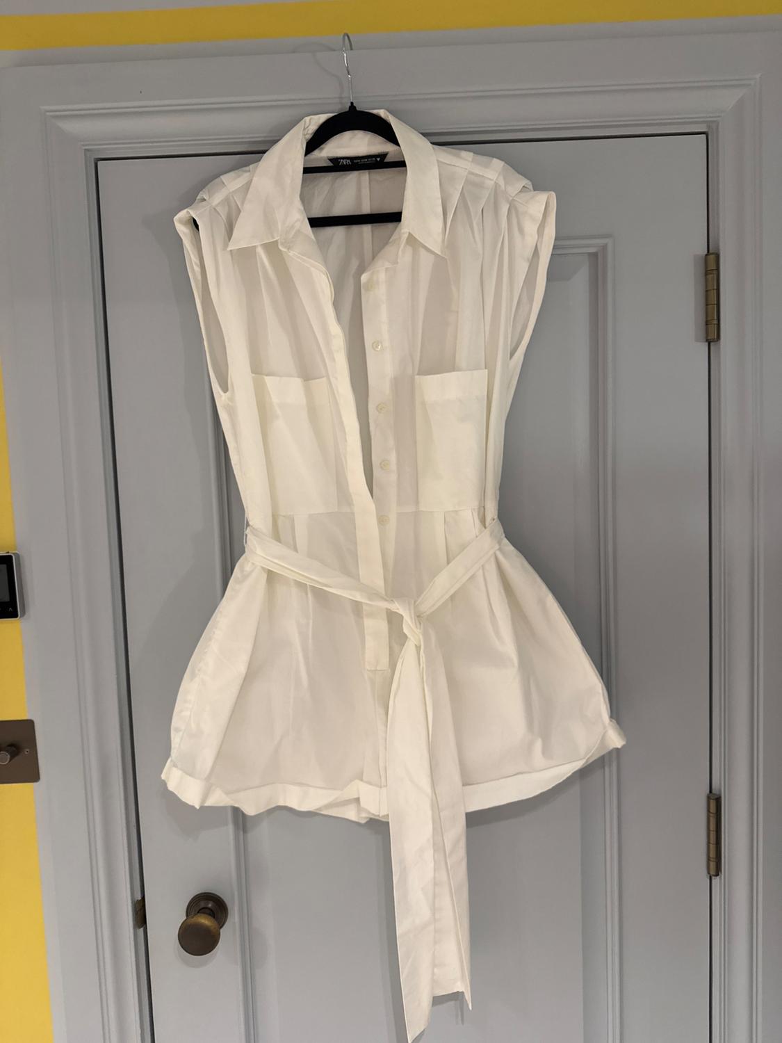 Sleeveless white blouse with belt, hanging on a grey door. Indoor setting with a yellow wall.