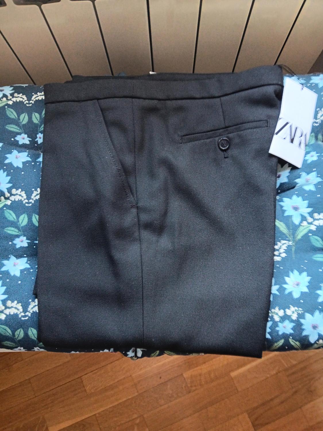Black trousers folded on a blue floral surface. Label with the text ZARA. Background with radiator and wooden floor.