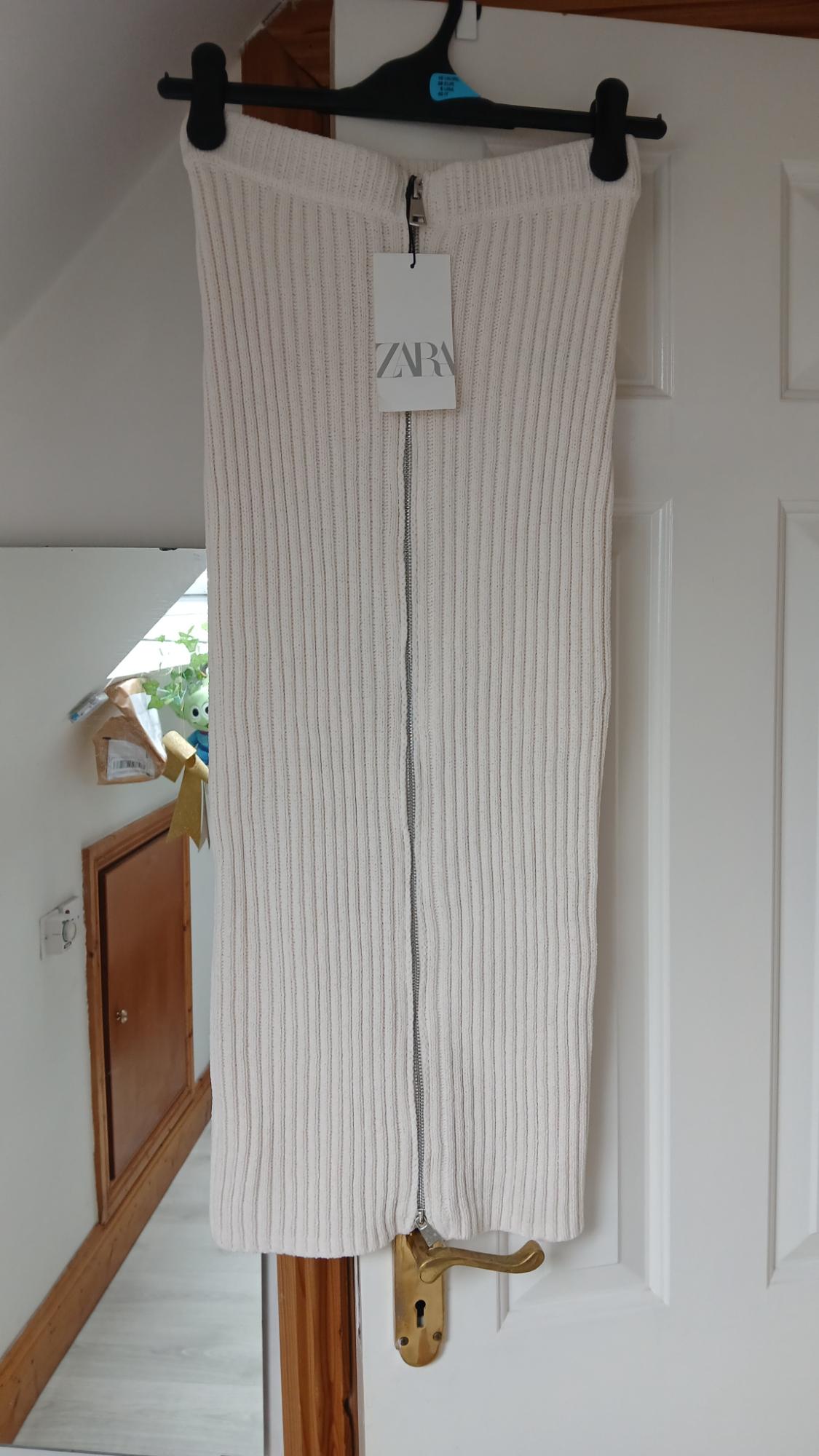 White ribbed knit skirt with a front zip hanging on a door. Label with ZARA text. Casual vibe.