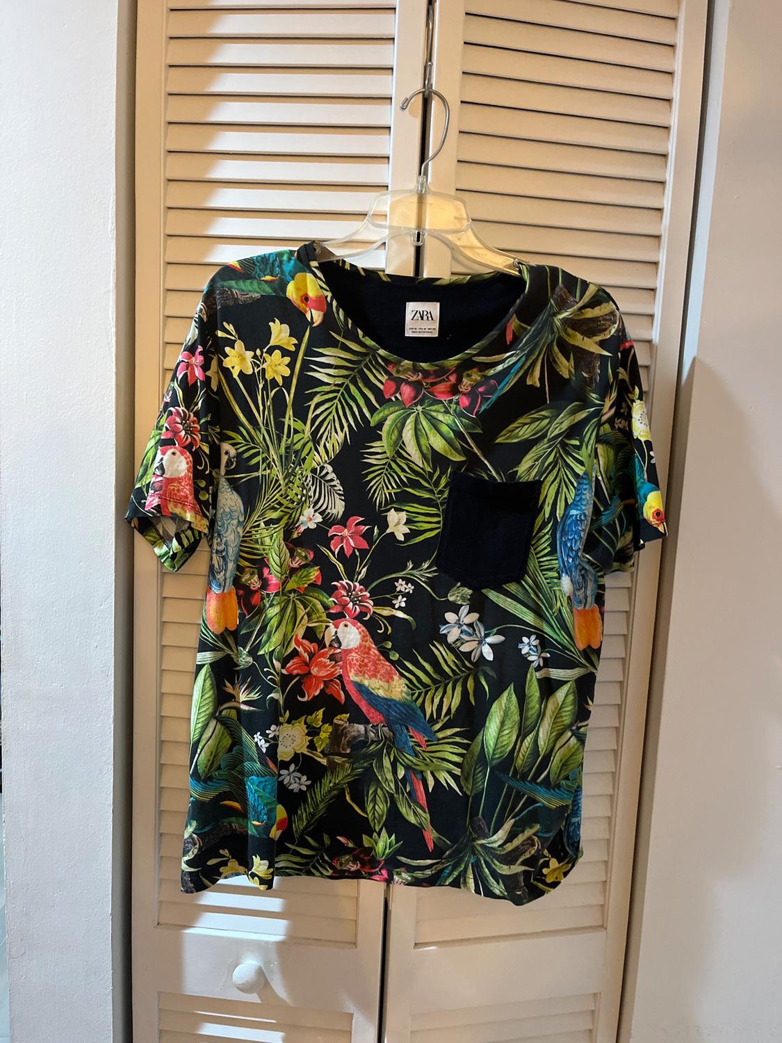 Hanging T-shirt with a tropical print of leaves and parrots in vibrant colours. Black background. Visible label: "Zara". Louvred door behind.