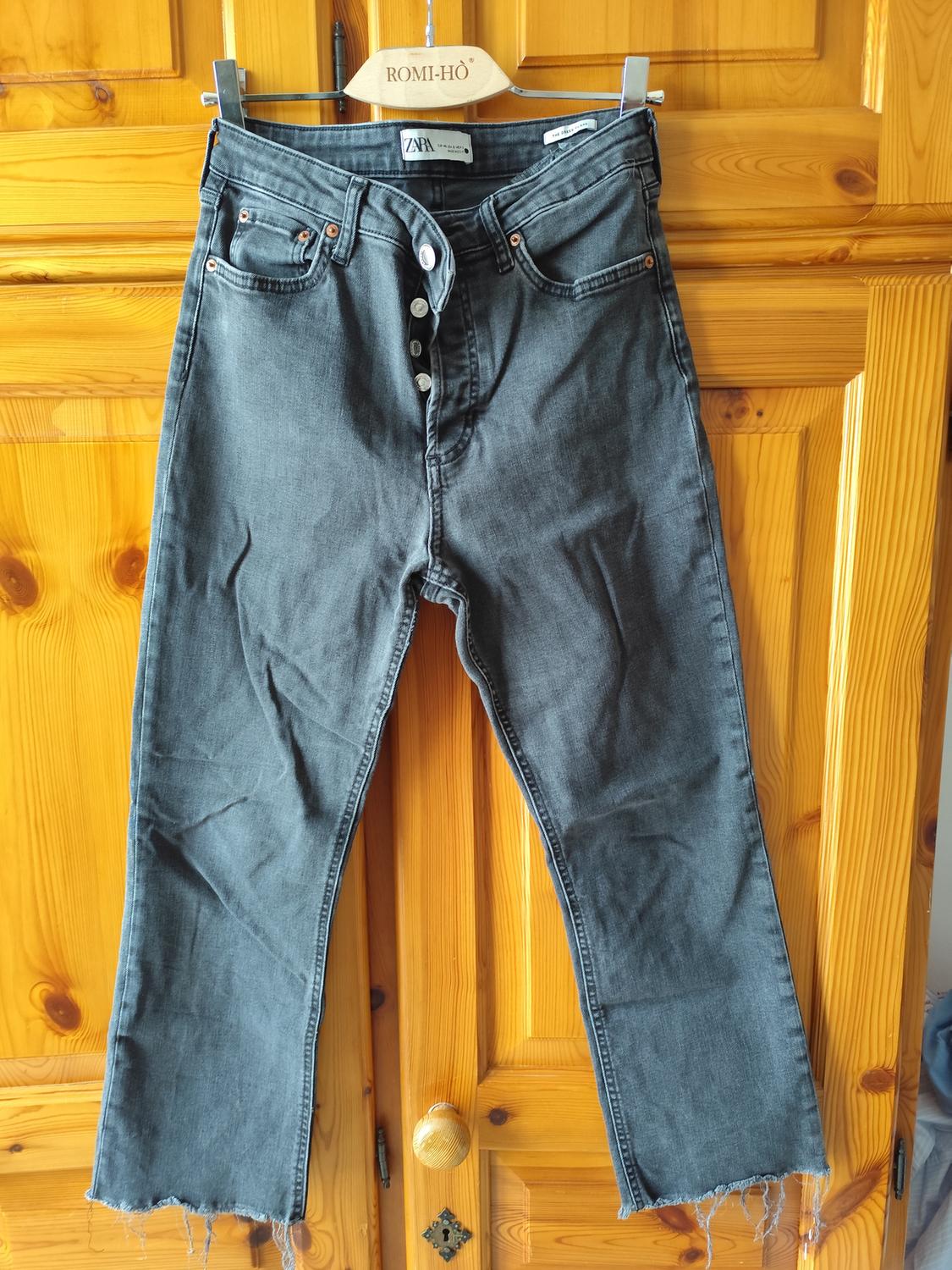 Worn grey jeans with visible buttons, hung on a wooden hanger in front of a light wood wardrobe door.
