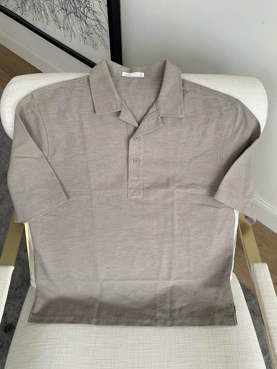 Beige short-sleeved polo shirt with ZARA text on the label, collar and buttons, on a white chair with gold armrests.