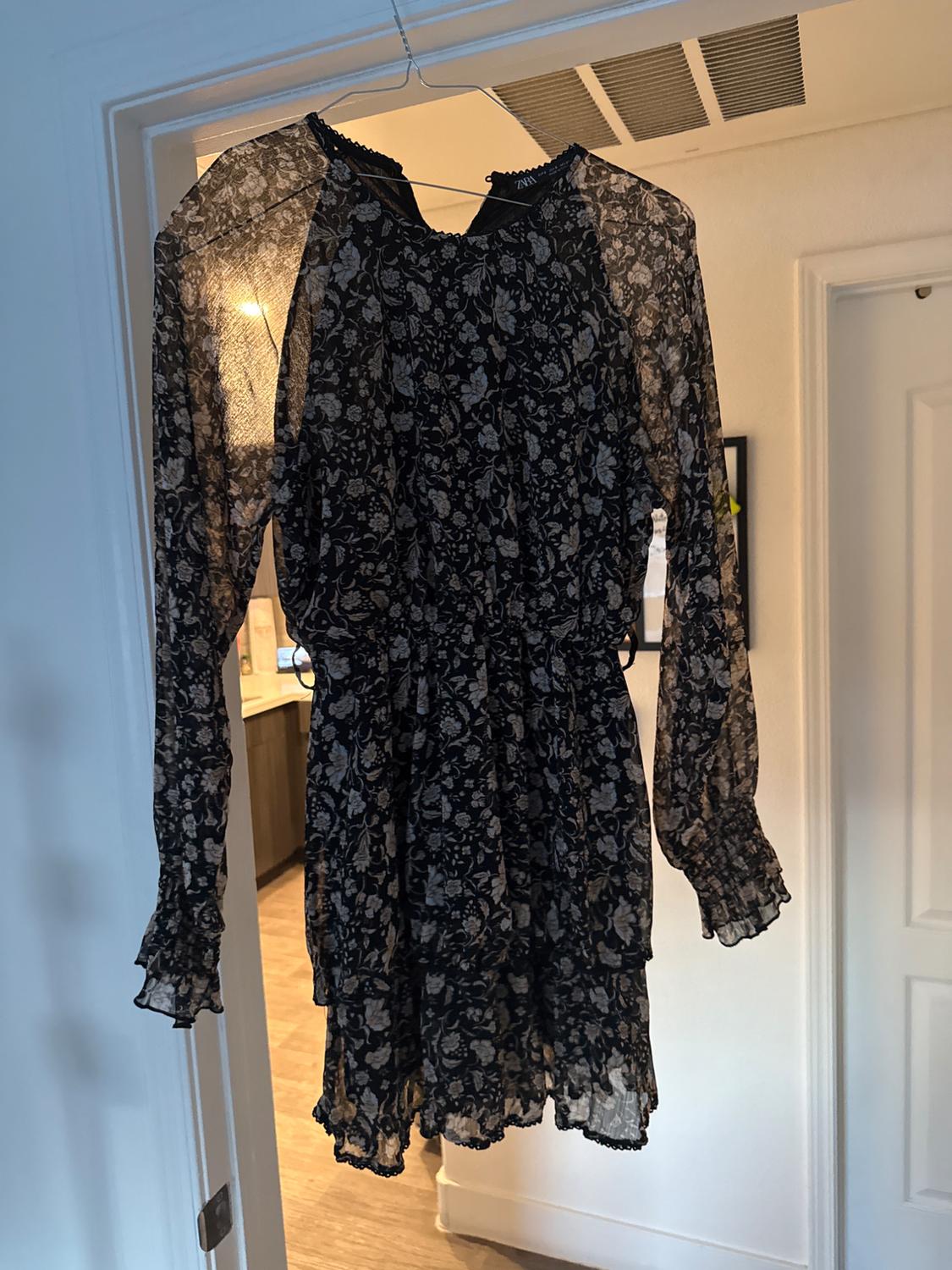 Short black dress with a white floral print, sheer long sleeves and frills, hanging on a door. ZARA text.