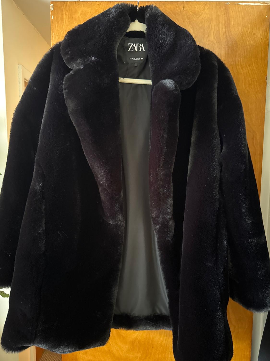 Black faux fur coat hanging on a white hanger with "ZARA" label visible, over a light wooden door.