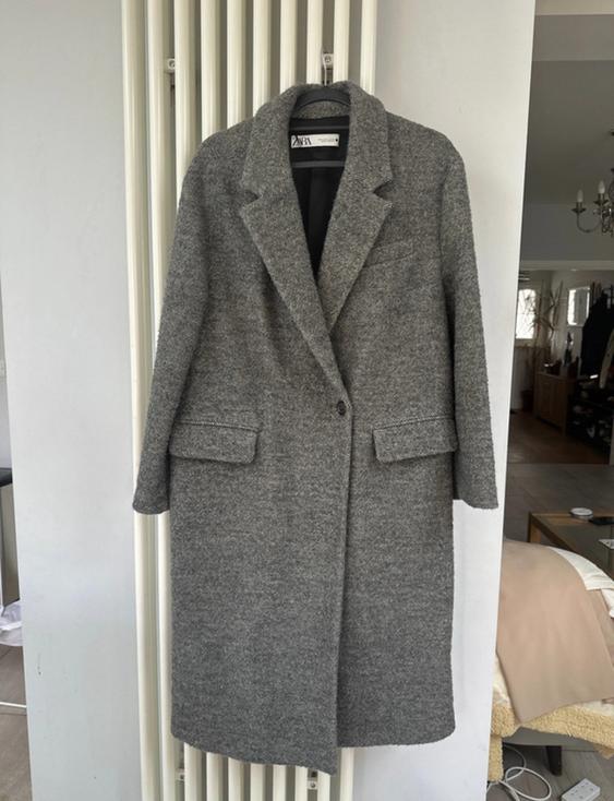 Grey wool coat hanging on a white radiator, with lapels and pockets. Living room background with furniture and chandelier.