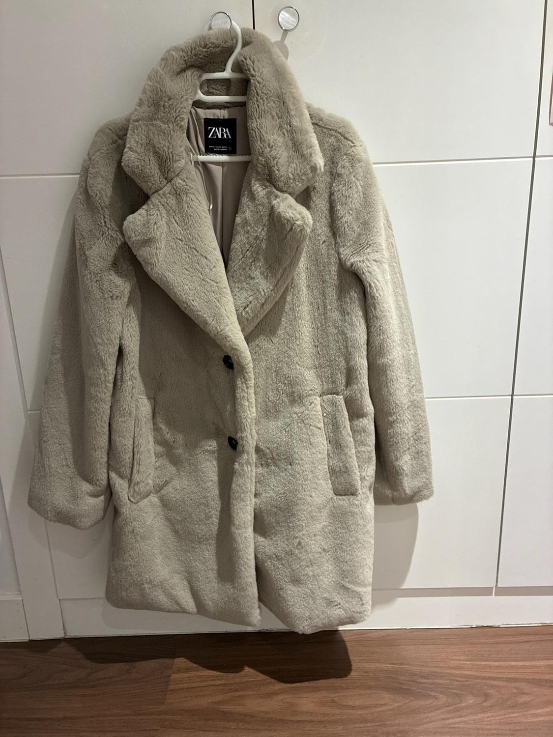 Beige faux-fur coat with lapels and dark buttons, hanging on a coat hanger over a white door. Interior label with the text ZARA.