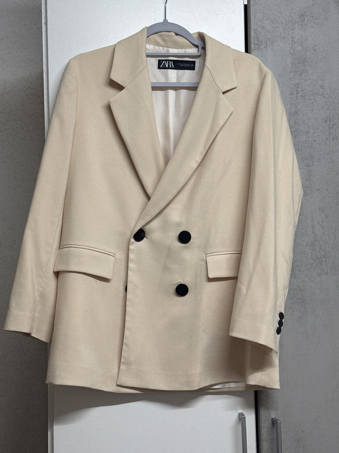 Beige double-breasted jacket with black buttons, hanging on a grey hanger. Visible label with the ZARA brand.